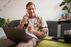 beeld van vent roeping iemand, Holding credit kaart en gebruik makend van laptop, heeft koffer en camera Ingepakt voor vakantie foto