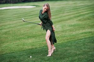 mooie vrouw gekleed in het groen staande op de golfbaan met stok in haar hand poseren voor de foto