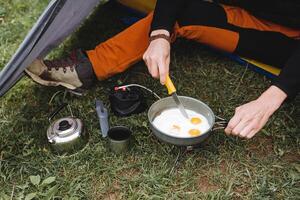 een toerist Aan een wandeltocht met een mes bezuinigingen door elkaar gegooid eieren, een vent kookt voedsel Aan een toerist brander, gebakken glazuur in een frituren pan in natuur, een toerist keuken camping. foto