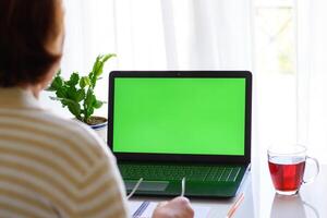 terug visie van senior vrouw vervelend bril, aan het kijken een laptop met groen scherm. ideaal voor presentatie van technologie gebruik door ouderen, afgelegen werk, online aan het leren foto