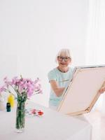 senior vrolijke vrouw kunstenaar in glazen met grijze haren schilderen bloemen in vaas. creativiteit, kunst, hobby, bezettingsconcept