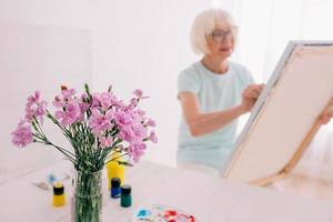 senior vrolijke vrouw kunstenaar in glazen met grijze haren schilderen bloemen in vaas. creativiteit, kunst, hobby, bezettingsconcept