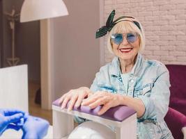 manicure meester in blauwe handschoenen afroming handen van oudere stijlvolle vrouw in blauwe zonnebril en spijkerjasje zittend op manicure salon