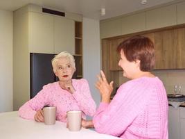 twee stijlvolle senior vrouwen in roze truien zitten met mokken op moderne keuken roddelen. vriendschap, praten, roddels, relaties, nieuws, familieconcept foto