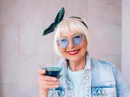 senior stijlvolle vrouw met grijs haar en in blauwe bril en spijkerjasje met glas met blauwe cocktail. alcohol, ontspanning, vakantie, pensioenconcept foto