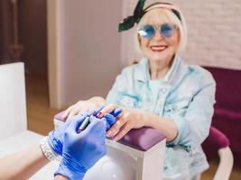 manicure meester in blauwe handschoenen afroming handen van oudere stijlvolle vrouw in blauwe zonnebril en spijkerjasje zittend op manicure salon