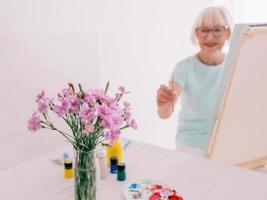 senior vrolijke vrouw kunstenaar in glazen met grijze haren schilderen bloemen in vaas. creativiteit, kunst, hobby, bezettingsconcept foto