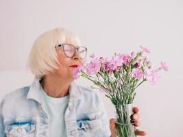 senior stijlvolle vrouw in bril en jeans jasje geur anjer bloemen in vaas. bloemen, pensioen, beroep, hobby concept foto