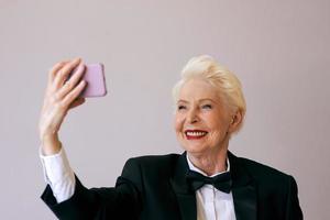 stijlvolle volwassen senior vrouw in smoking met mobiel videobellen of selfie maken. plezier, feest, stijl, levensstijl, bedrijf, technologie, feestconcept foto