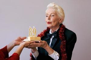 gelukkige vrolijke stijlvolle negenentachtig jaar oude vrouw in zwart pak viert haar verjaardag met taart. levensstijl, positief, mode, stijlconcept foto