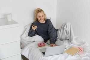 portret van glimlachen openhartig vrouw, aan het liegen in bed met donut, gebruik makend van smartphone en laptop, resting Bij huis in slaapkamer, aan het kijken TV tonen of chatten online foto