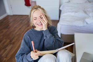 portret van charismatisch blond meisje, glimlachen vrouw in slaapkamer, Holding notitieboekje en pen, schrijven in logboek of dagboek, creëert naar Doen lijst in ontwerper foto