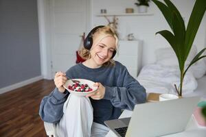 portret van glimlachen mooi vrouw, zittend in kamer met ontbijt, aan het eten en aan het kijken TV tonen Aan laptop, lachend en op zoek Bij scherm foto