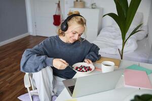 portret van glimlachen jong vrouw, aan het kijken TV tonen in koptelefoon, aan het eten ontbijt en op zoek Bij laptop scherm foto