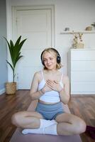 verticaal schot van jong sportief vrouw in koptelefoon, zit Aan rubber mat na training, Holding handen Aan borst, looks terzijde met vredevol, ontspannen gezicht uitdrukking foto