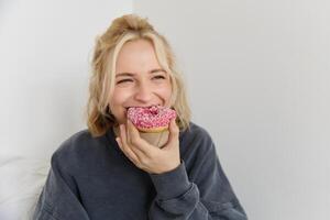 dichtbij omhoog portret van Vrolijk, schattig blond vrouw, Holding donut, aan het eten zoet, heerlijk comfort voedsel, tonen toetje Bij camera foto