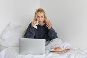 portret van verdrietig en depressief vrouw, gevoel diepbedroefd, aan het liegen in bed met comfort voedsel, aan het eten donut, huilen van verontrustend film, aan het kijken iets Aan laptop foto