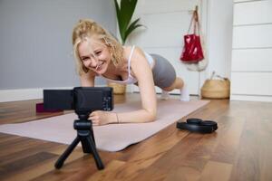 portret van jong sportief vlogger, geschiktheid instructeur staand in plank Aan rubber yoga mat, opname van haarzelf aan het doen training Bij huis foto