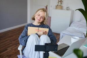 portret van blond glimlachen vrouw, records Aan digitaal camera hoe ze schrijft in notitieboekje, praat naar volgers, aan het doen levensstijl blog inhoud in haar kamer foto