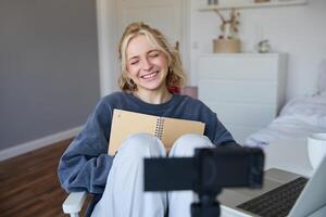 portret van blond glimlachen vrouw, records Aan digitaal camera hoe ze schrijft in notitieboekje, praat naar volgers, aan het doen levensstijl blog inhoud in haar kamer foto