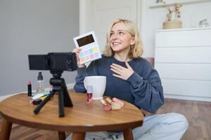 portret van mooi levensstijl blogger, meisje records een Aan haar camera voor sociaal media, shows palet van oogschaduw, doet een bedenken zelfstudie voor haar volgers, zit in haar kamer foto