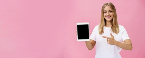 meisje beveelt aan gebruik enkel en alleen deze digitaal tablet. charismatisch jong vrouw met eerlijk haar- gebruind huid en Nee bedenken in wit t-shirt richten Bij apparaatje scherm en glimlachen vriendelijk Bij camera over- roze muur foto