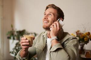 portret van knap jong Mens pratend Aan de telefoon, zittend in cafe en drinken koffie, beantwoorden een telefoontje foto