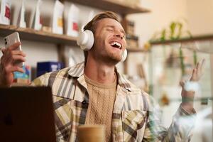 portret van knap gelukkig jongen, freelancer werken en luisteren naar muziek- in draadloze koptelefoon, dansen Aan zijn stoel in cafe, genieten van favoriete lied in oortelefoons foto