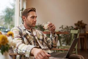 portret van jong freelancer, een Mens in gewoontjes kleren, zittend in cafe en drinken koffie, werken van een afstand Aan laptop, digitaal nomade aan het doen zijn project buitenshuis in samenwerken ruimte foto