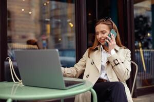 glimlachen Europese zakenvrouw pratend telefoon terwijl werken online zittend Bij buitenshuis cafe terras foto