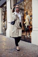 elegant vrouw in zonnebril is pratend telefoon Aan stad straat achtergrond en op zoek Bij kant foto