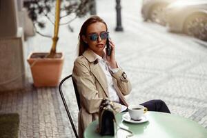 mooi vrouw vervelend zonnebril zittend Bij cafe terras pratend Aan telefoon terwijl drinken koffie foto