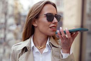 gelukkig zakenvrouw in zonnebril is opname audio bericht Aan telefoon Aan stad straat achtergrond foto