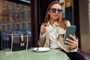 glimlachen elegant vrouw in bril zittend Bij cafe terras en Holding telefoon terwijl drinken koffie foto