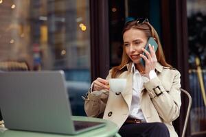 aantrekkelijk Europese vrouw pratend telefoon terwijl werken online zittend Bij buitenshuis cafe terras foto