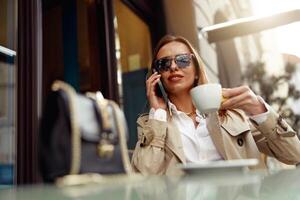glimlachen vrouw vervelend zonnebril zittend Bij cafe terras pratend Aan telefoon terwijl drinken koffie foto