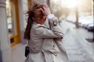 glimlachen aantrekkelijk vrouw in jas kruispunt de stad straat en op zoek Bij camera. wazig achtergrond foto