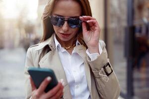 modieus Europese vrouw in zonnebril is op zoek Aan telefoon Aan stad straat achtergrond foto