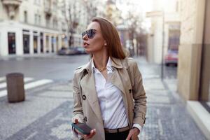 aantrekkelijk Europese vrouw in zonnebril is staand met telefoon Aan stad straat achtergrond foto