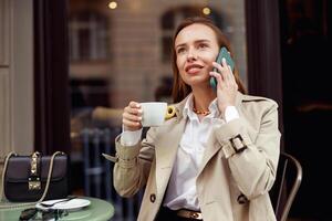 glimlachen vrouw zittend Bij cafe terras en pratend Aan telefoon terwijl drinken koffie foto