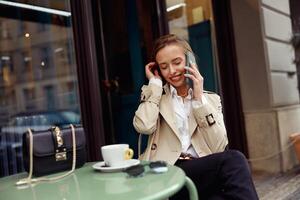 glimlachen elegant vrouw zittend Bij cafe terras pratend Aan telefoon terwijl drinken koffie foto
