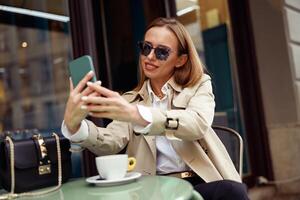 glimlachen elegant vrouw in bril zittend Bij cafe terras en nemen selfie Aan telefoon foto