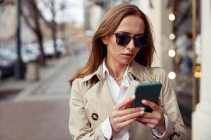 aantrekkelijk Europese vrouw in zonnebril is staand met telefoon Aan stad straat achtergrond foto