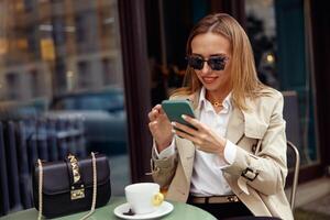 glimlachen elegant vrouw in bril zittend Bij cafe terras en gebruik telefoon terwijl drinken koffie foto