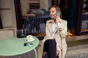 glimlachen elegant vrouw zittend Bij cafe terras pratend Aan telefoon terwijl drinken koffie foto