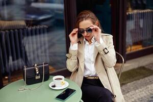 elegant jong vrouw vervelend zonnebril terwijl drinken koffie Aan restaurant terras foto