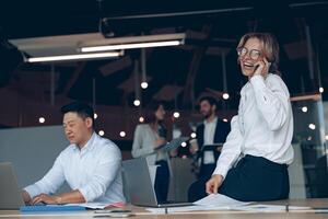 glimlachen volwassen bedrijf vrouw zittend Aan tafel en pratend telefoon gedurende vergadering in modern kantoor foto