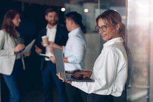 volwassen glimlachen bedrijf dame Bij kantoor met groep van collega's Aan achtergrond, werken Aan laptop foto