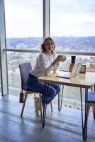 midden- leeftijd vrouw met blond haar- drinken koffie en werken Aan een laptop in een coworking ruimte met panoramisch ramen en een visie van de stad van bovenstaand foto
