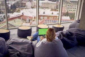 een vrouw in gewoontjes kleren is zittend Bij een laptop Aan een poef in voorkant van een panoramisch venster met een visie van de stad, groen scherm, chroma sleutel foto